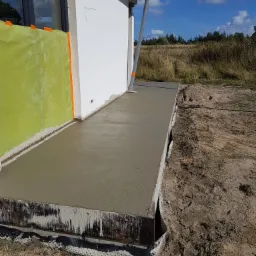 Wylewka betonowa, świeżo położona, przy ścianie budynku z oknem, widoczna folia ochronna i rynna spustowa, tło to łąka i niebo.