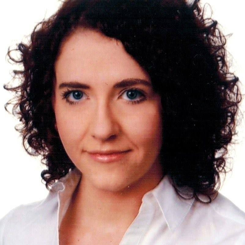 Portret kobiety o kręconych, ciemnych włosach i jasnych, niebieskich oczach, ubrana w białą koszulę.