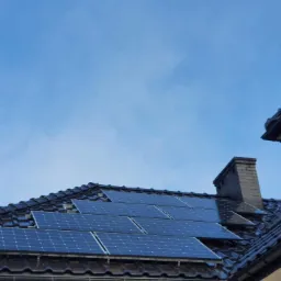 Panele słoneczne na dachu z ciemnej dachówki, widoczne mocowania i komin z cegły na tle błękitnego nieba.