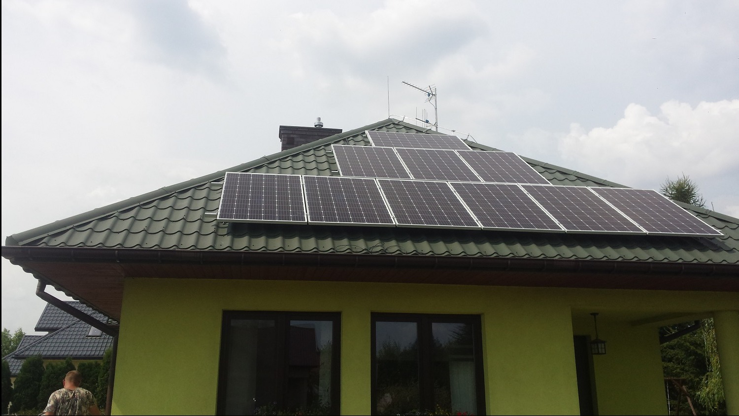 Panele słoneczne zamontowane na zielonym, spadzistym dachu domu jednorodzinnego z zieloną elewacją. Widoczny komin, antena oraz fragment sąsiedniego domu.