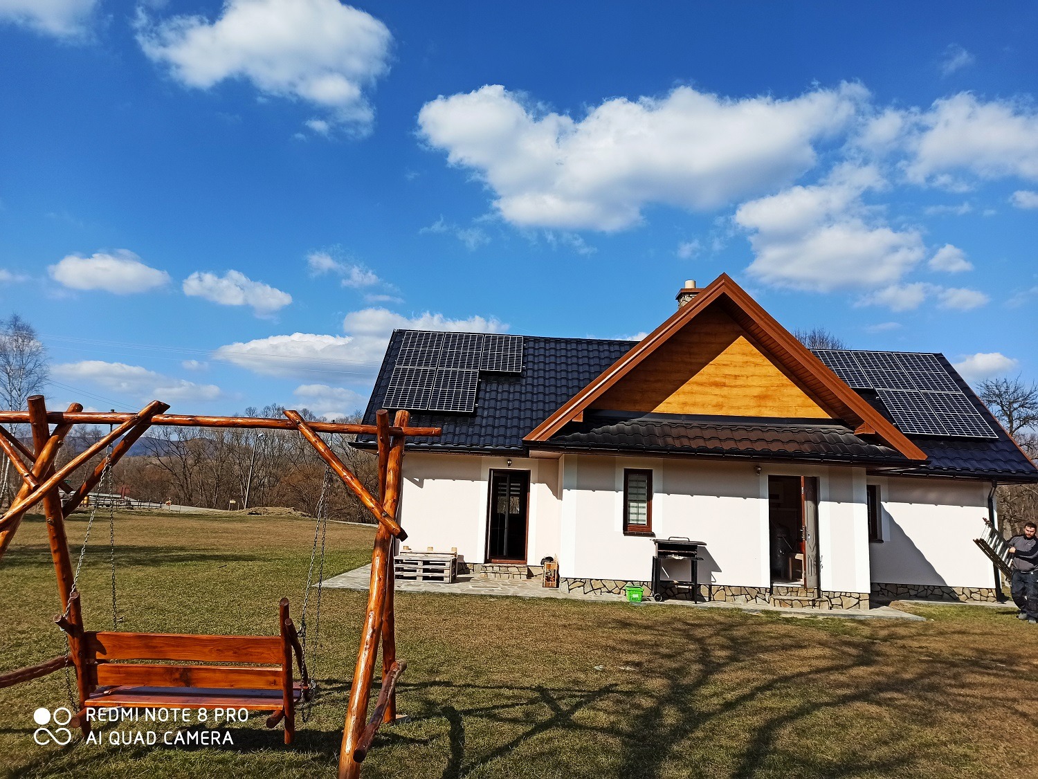 Dom jednorodzinny z panelami słonecznymi na dachu, widok z ogrodu z drewnianą huśtawką, błękitne niebo z chmurami w tle.