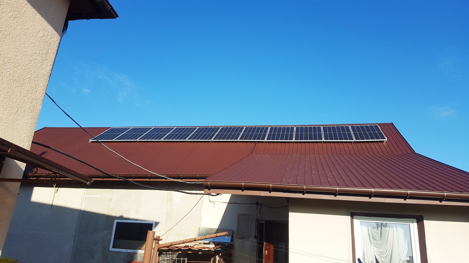 Panele słoneczne zamontowane na brązowym dachu domu jednorodzinnego, widoczne kable i orynnowanie, błękitne niebo w tle.