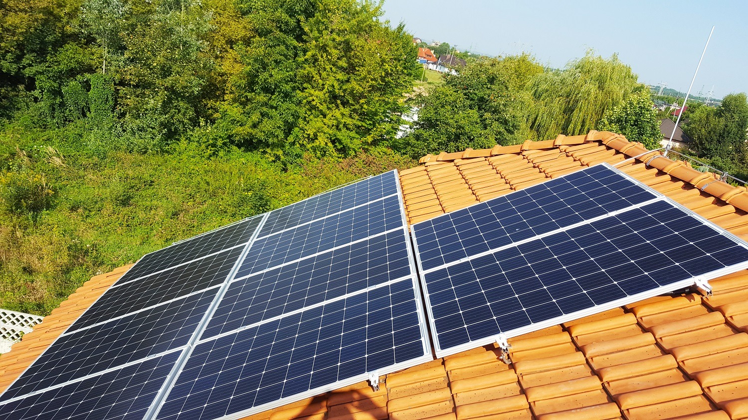 Panele fotowoltaiczne zamontowane na pomarańczowej dachówce, widok z góry, w tle zieleń i zabudowania