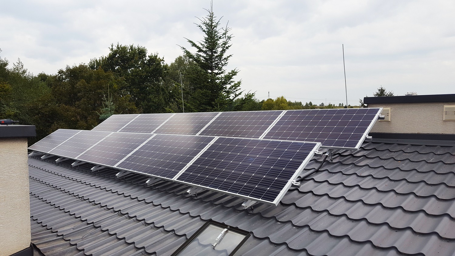 Panel słoneczny zamontowany na dachu z dachówki w odcieniach szarości, widok z góry, w tle drzewa i pochmurne niebo.