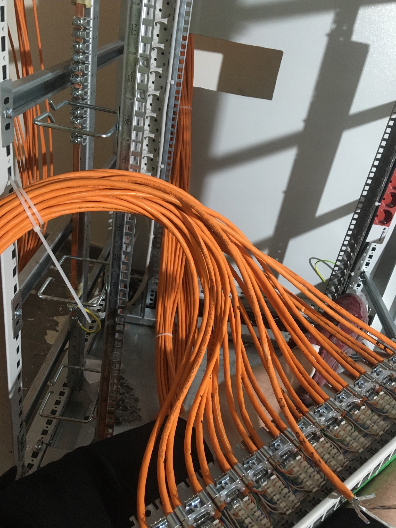 Pomarańczowe kable telekomunikacyjne podłączone do panelu krosowniczego w szafie rack, widoczne metalowe elementy mocujące i etykiety na kablach.