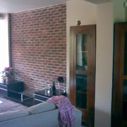 Salon z dekoracyjną ścianą z cegły, ciemnymi meblami i częściowo widoczną sofą, oświetlony przez okno po lewej stronie.