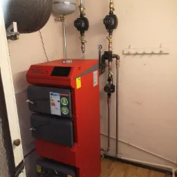 Czerwony piec CO Tekla Draco D Bio z szarymi drzwiczkami, podłączony do instalacji z pompami obiegowymi i zbiornikiem wyrównawczym w kotłowni z drewnianą podłogą.