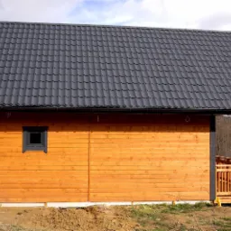 Nowo wybudowany, parterowy budynek z drewnianych bali, z ciemną dachówką i małym oknem, z zadaszonym tarasem, częściowo posadowiony na bloczkach betonowych.