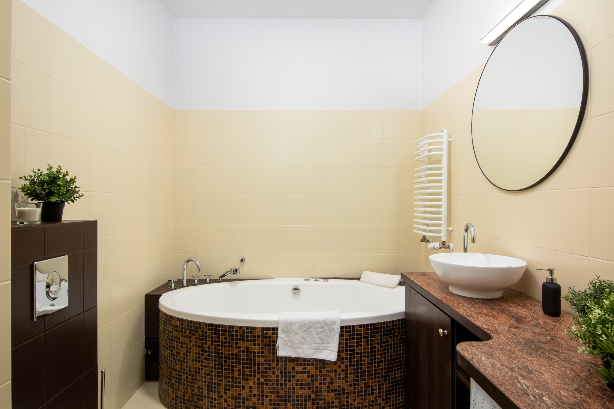 Nowoczesna łazienka z owalną wanną obłożoną mozaiką, umywalką nablatową i beżowymi płytkami ściennymi.