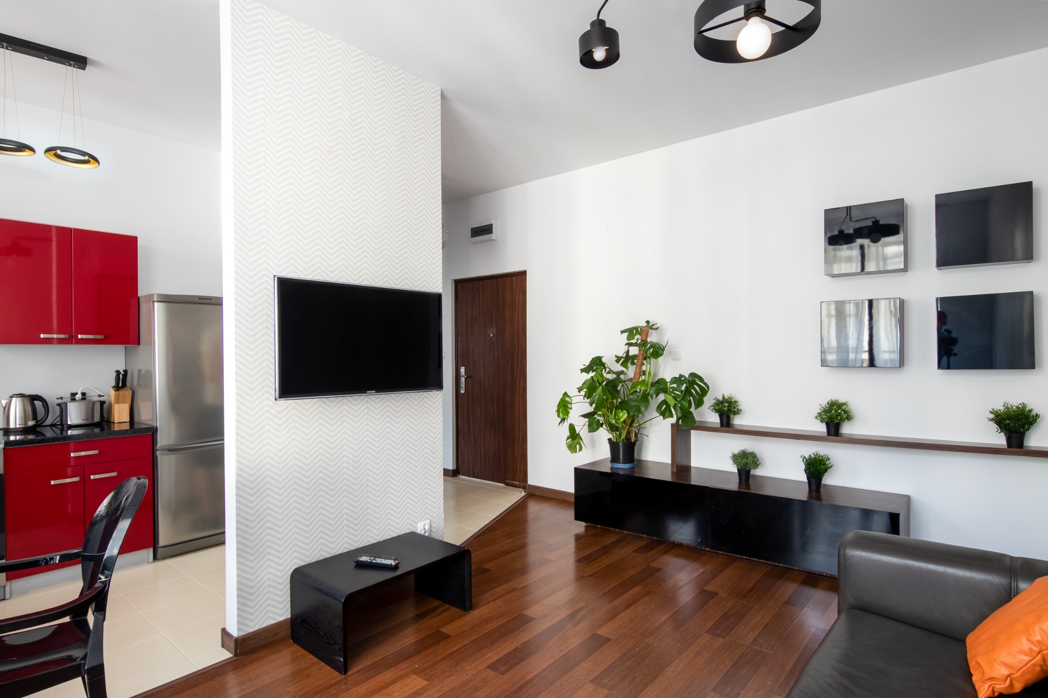 Nowoczesny salon z drewnianą podłogą, telewizorem na ścianie w geometryczny wzór, czarnymi meblami, roślinami doniczkowymi i częściowo widoczną kuchnią z czerwonymi szafkami.