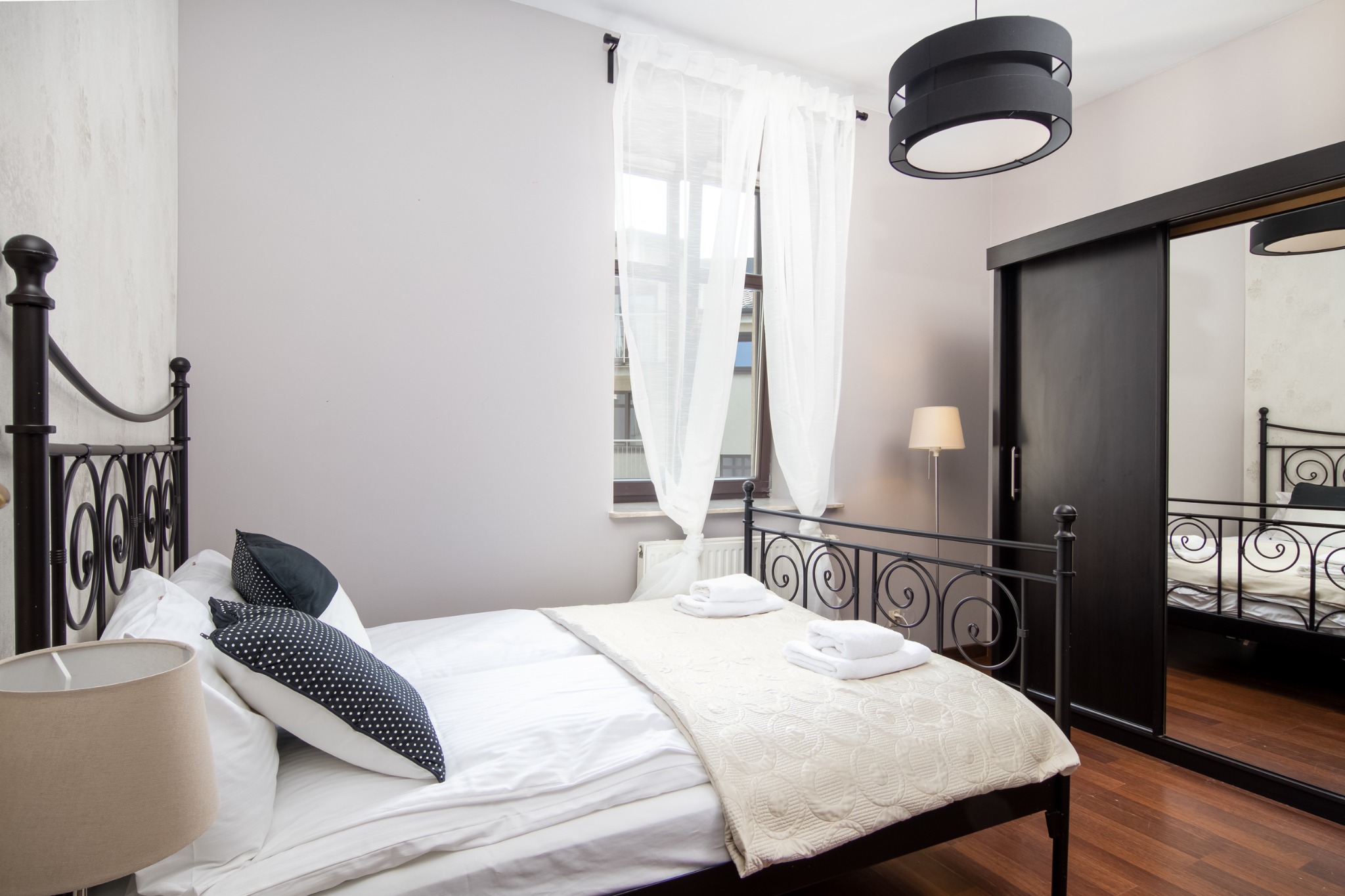 Jasne wnętrze sypialni z czarnym, metalowym łóżkiem, pościelą w odcieniach bieli i beżu, oraz czarną szafą przesuwną z lustrem, oświetloną lampą podłogową i wiszącą.