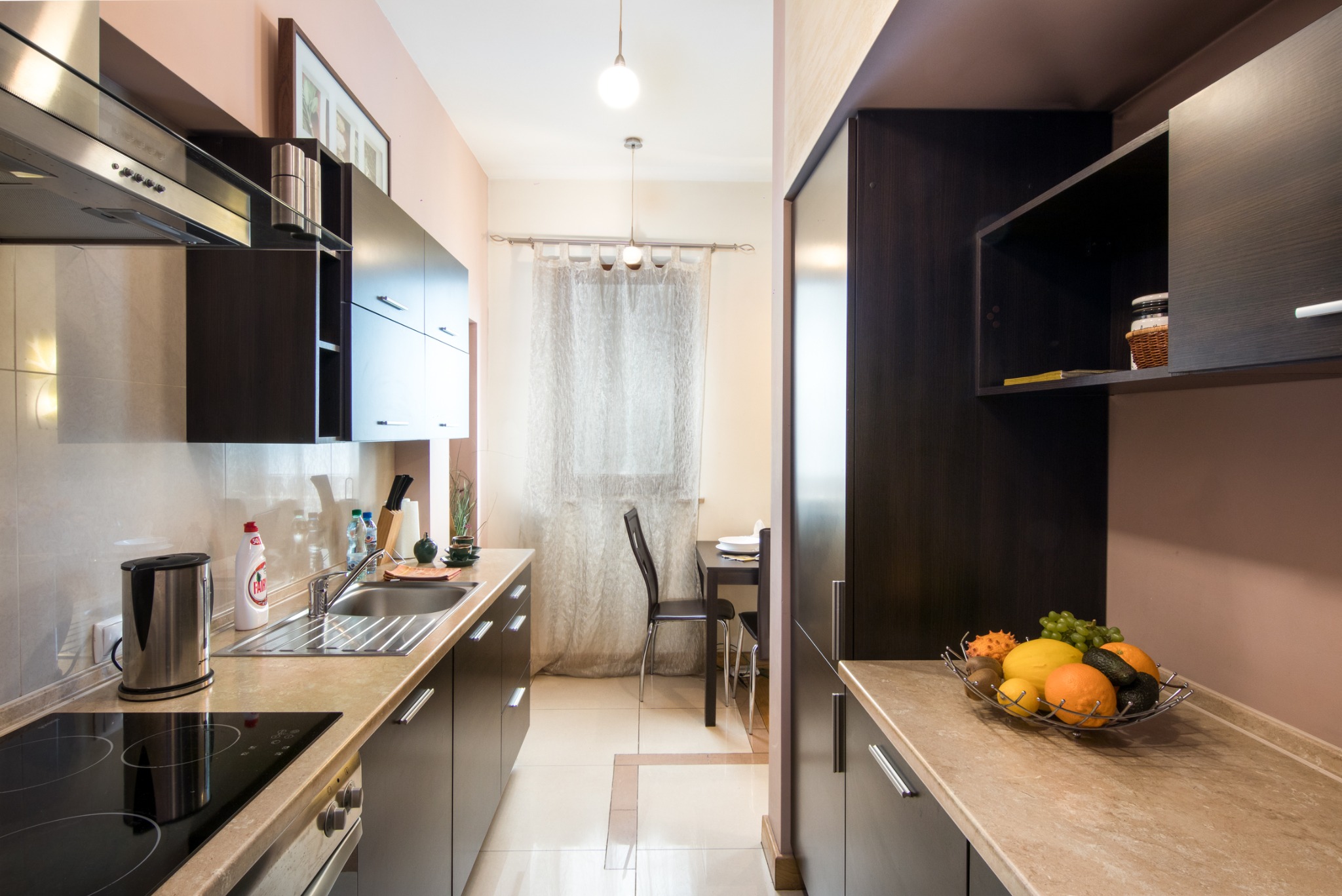 Nowoczesna kuchnia w beżowo-brązowych odcieniach z ciemnymi szafkami, jasnym blatem i owocami w metalowej misie, widok w głąb pomieszczenia na stół z krzesłami.