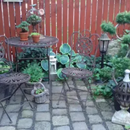 Rustykowy zestaw mebli ogrodowych z kutego żelaza, składający się z okrągłego stolika i dwóch krzeseł, otoczony różnorodną zielenią, w tym roślinami w doniczkach i pnączami, na tle czerwonego...