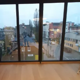 Duże okno z czarnymi ramami, widok na miasto z budynkami, drzewami i ulicą w pochmurny dzień, jasna drewniana podłoga w pomieszczeniu.