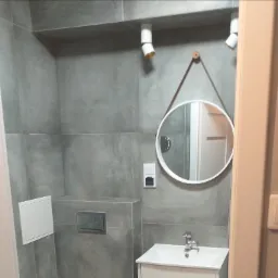 Nowoczesna łazienka z szarymi płytkami, okrągłym lustrem na skórzanym pasku i punktowym oświetleniem, widok zza futryny drzwi.