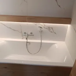 Biała wanna obudowana panelami imitującymi drewno, z chromowaną baterią i słuchawką prysznicową na tle marmurowej ściany z podświetleniem LED.