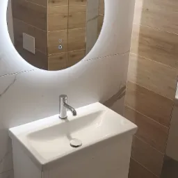 Nowoczesna łazienka z okrągłym lustrem podświetlanym LED, białą umywalką z szafką i armaturą chromowaną, wykończona płytkami imitującymi marmur i drewno.