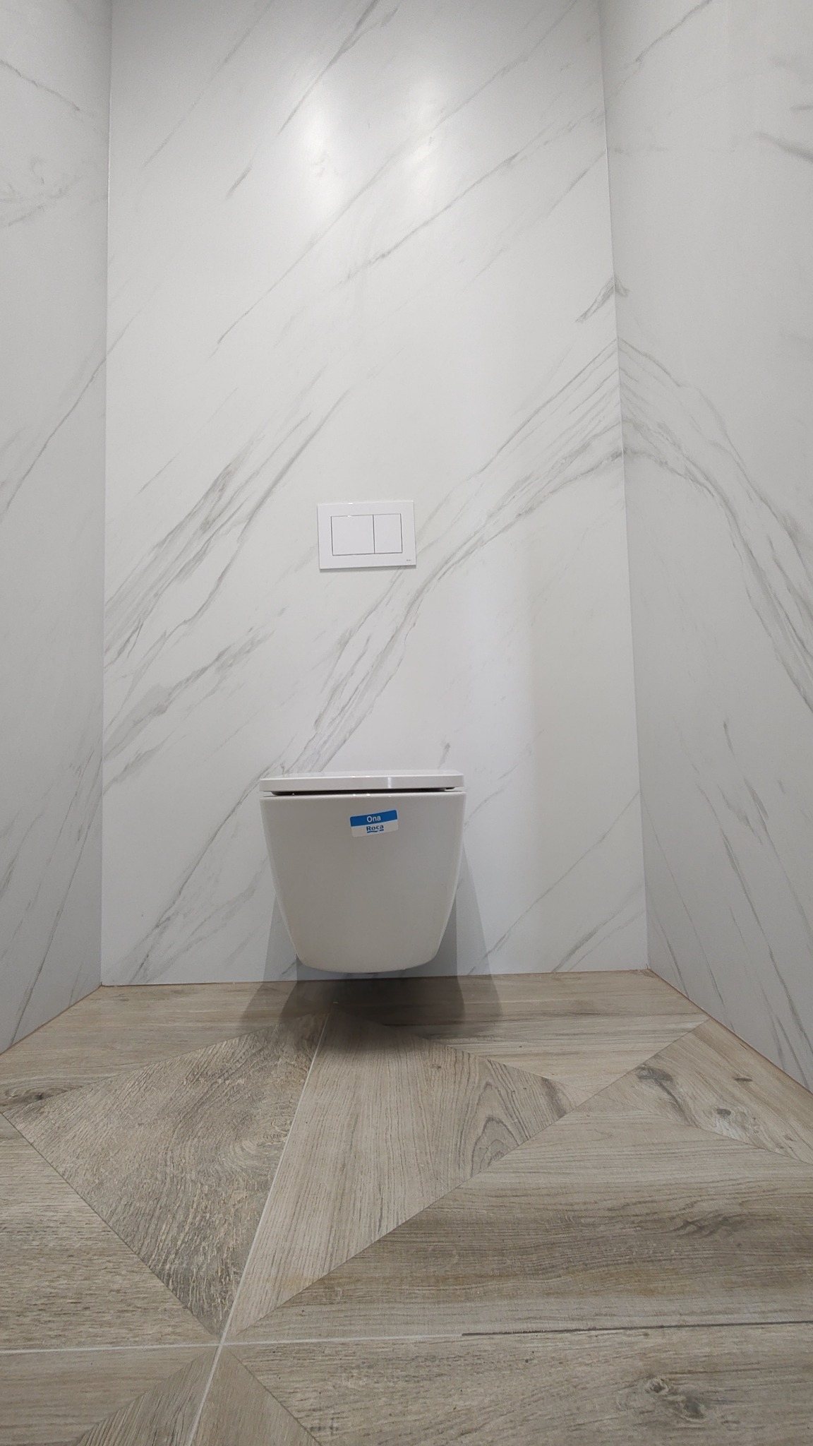 Nowoczesna łazienka z podwieszaną toaletą Roca Ona, wykończona płytkami imitującymi marmur na ścianach i drewnopodobnymi płytkami ułożonymi w geometryczny wzór na podłodze.