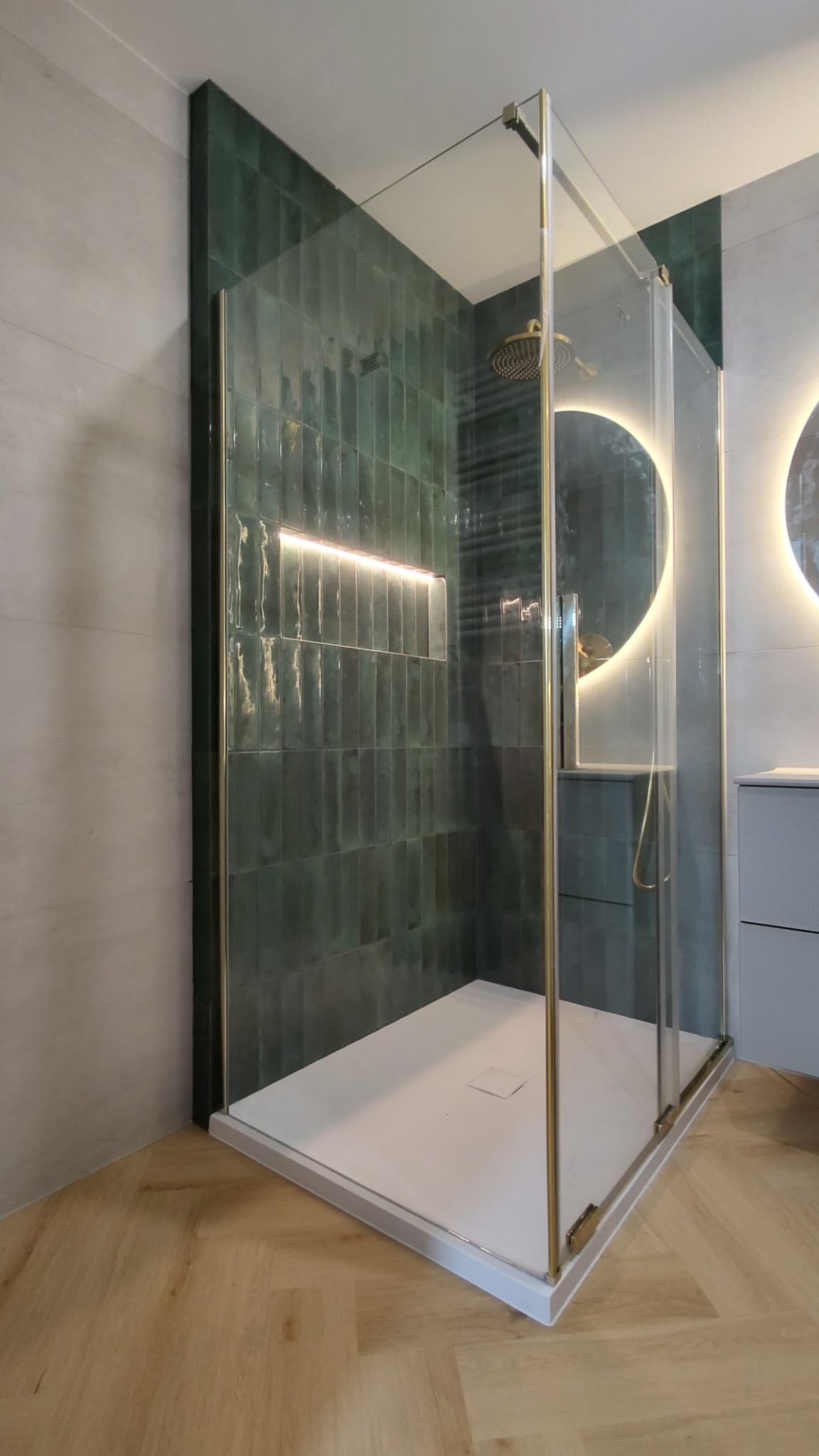 Kabina prysznicowa z zielonymi płytkami wertykalnymi, podświetlana wnęka, złote wykończenia, biały brodzik i drewnopodobna podłoga ułożona w jodełkę.