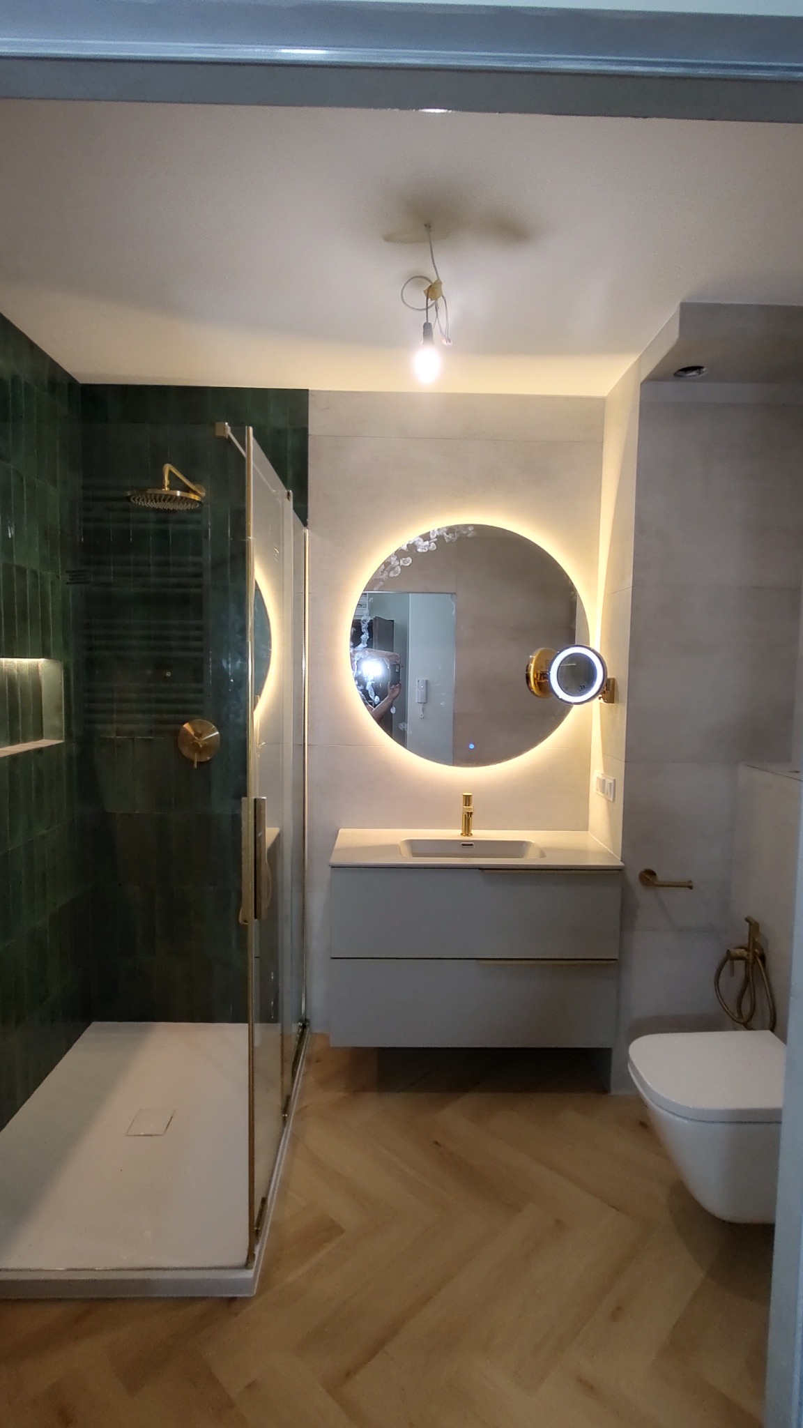 Nowoczesna łazienka z kabiną prysznicową typu walk-in z ciemnozielonymi płytkami, okrągłym lustrem z podświetleniem LED, umywalką nablatową z szafką w kolorze gołębim i toaletą w stylu...