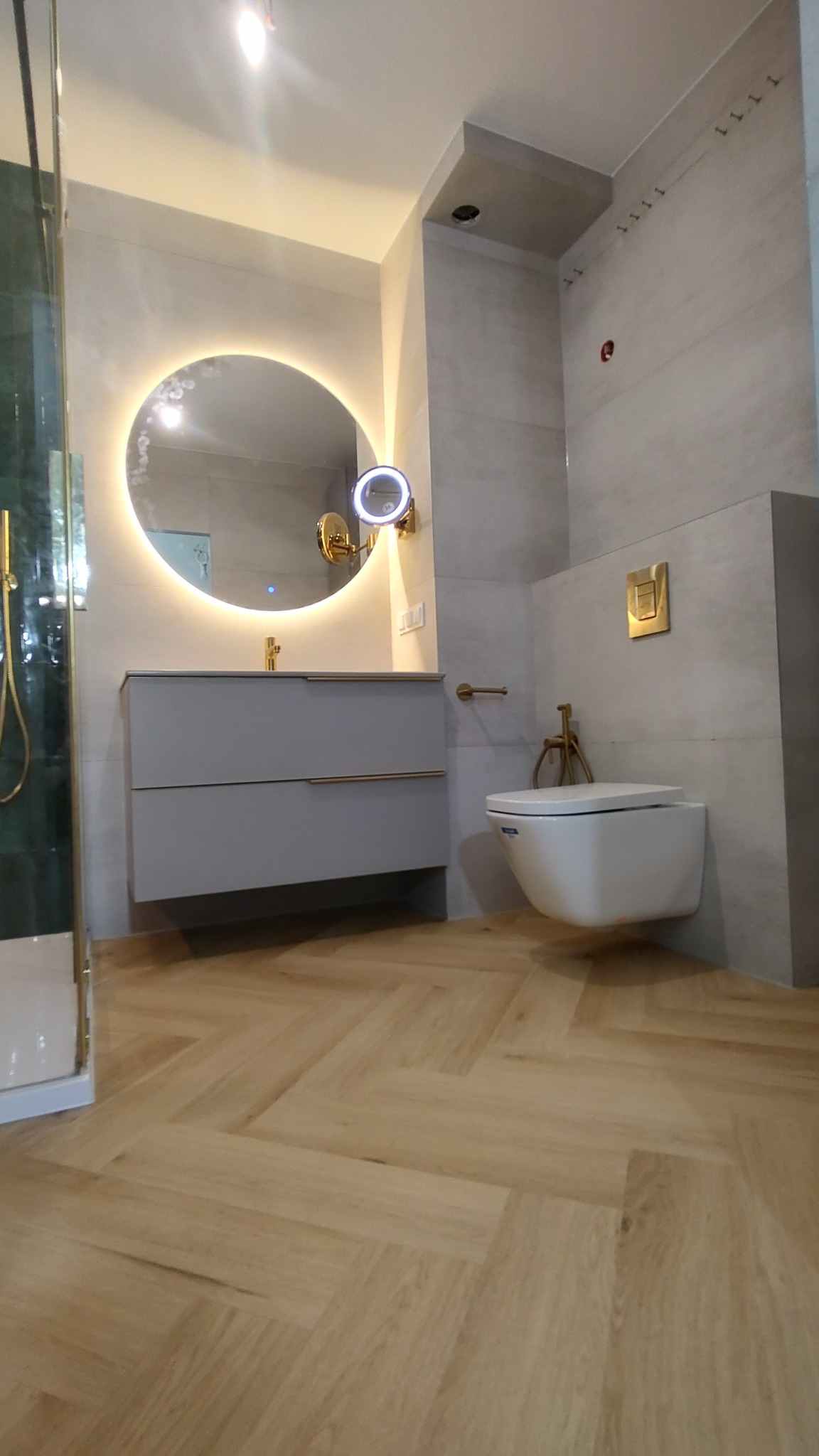 Nowoczesna łazienka z podwieszaną toaletą, umywalką z szafką w kolorze szarym, okrągłym lustrem z podświetleniem LED i podłogą ułożoną w jodełkę.