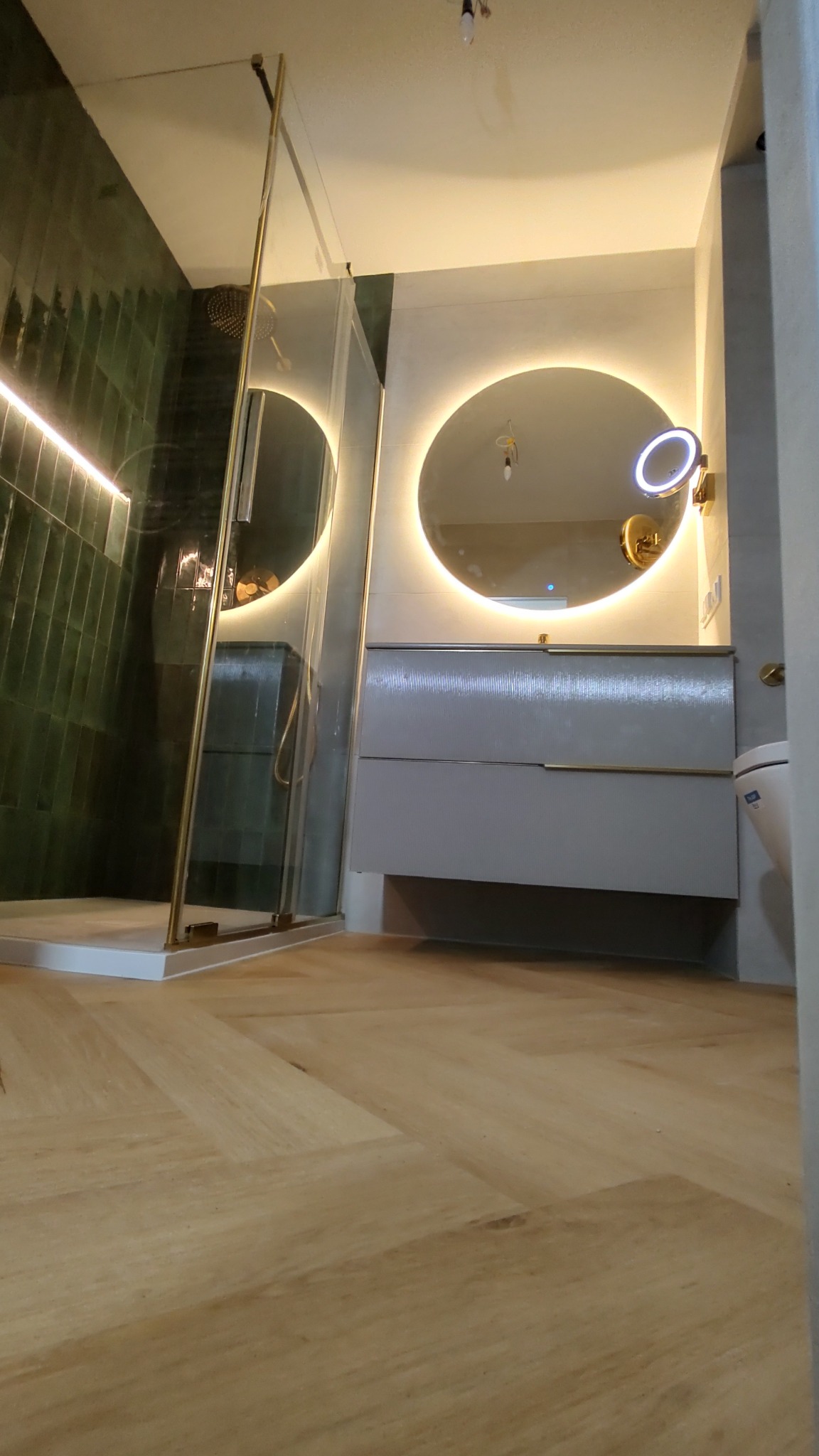 Nowoczesna łazienka z kabiną prysznicową typu walk-in z złotymi detalami, okrągłym lustrem z podświetleniem LED, szarą szafką pod umywalkę i drewnianą podłogą ułożoną w jodełkę.