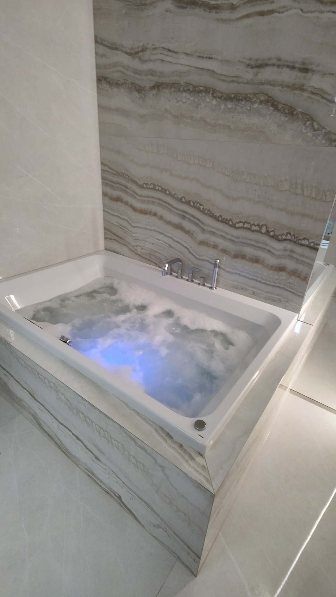 Wanna z hydromasażem wypełniona wodą, podświetloną na niebiesko, otoczona płytkami imitującymi marmur w łazience w Ciecierzycach.