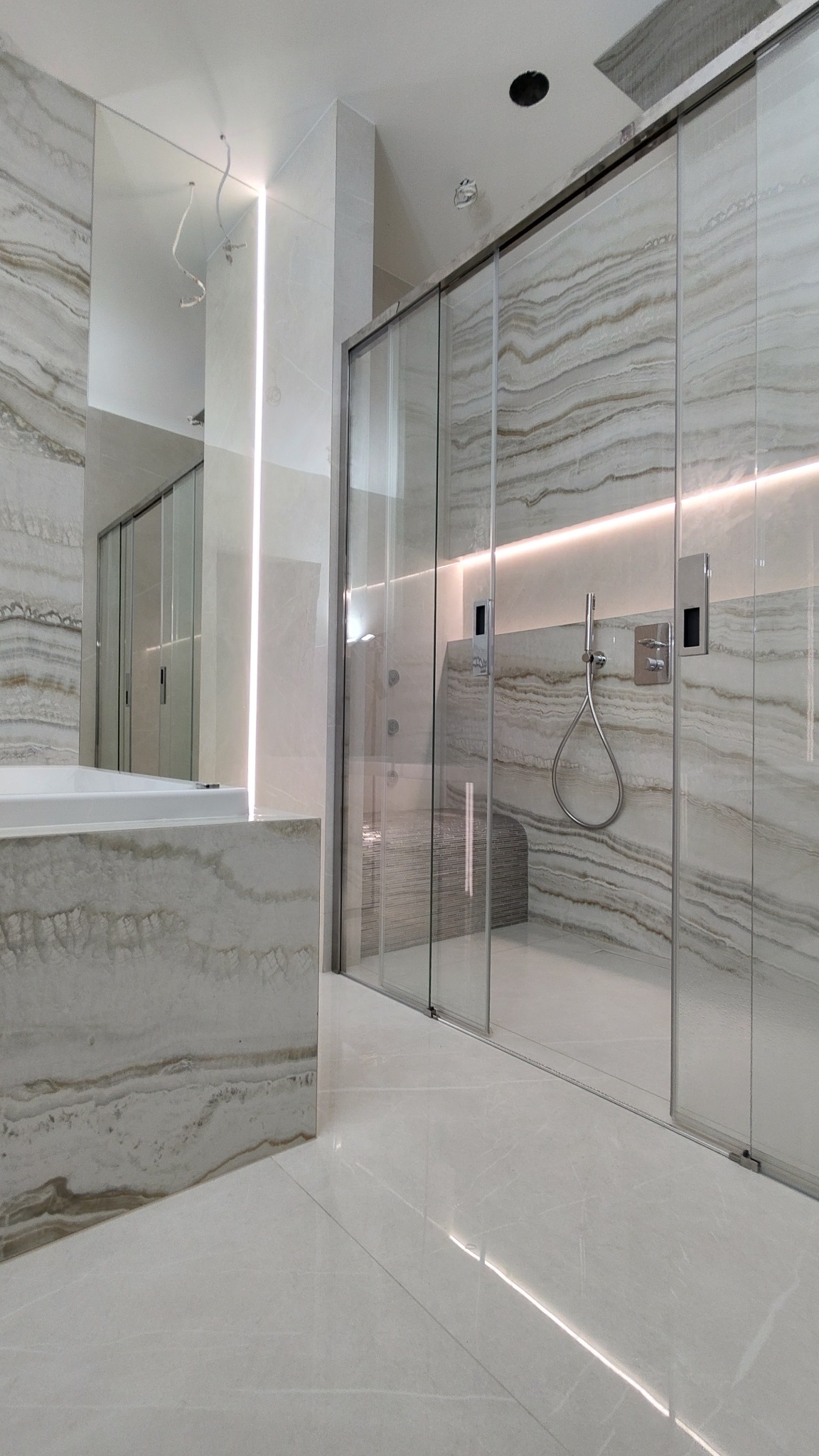 Nowoczesna łazienka z kabiną prysznicową typu walk-in, wykończona płytkami imitującymi marmur w odcieniach beżu i szarości, oświetlenie LED podkreślające fakturę płytek, minimalistyczna armatura.