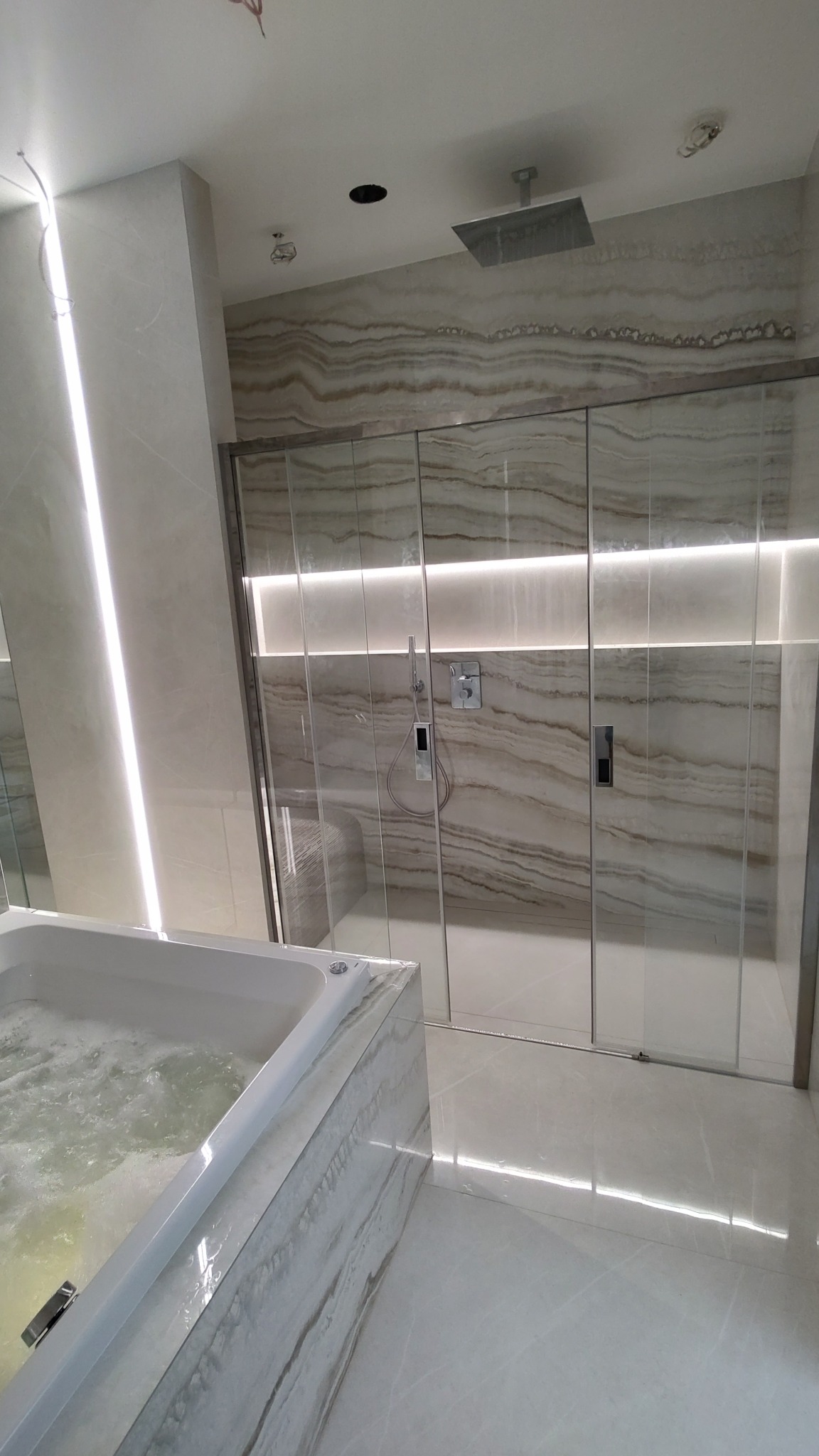 Nowoczesna łazienka z wanną z hydromasażem i kabiną prysznicową typu walk-in, wykończona płytkami imitującymi onyks z subtelnym podświetleniem LED.