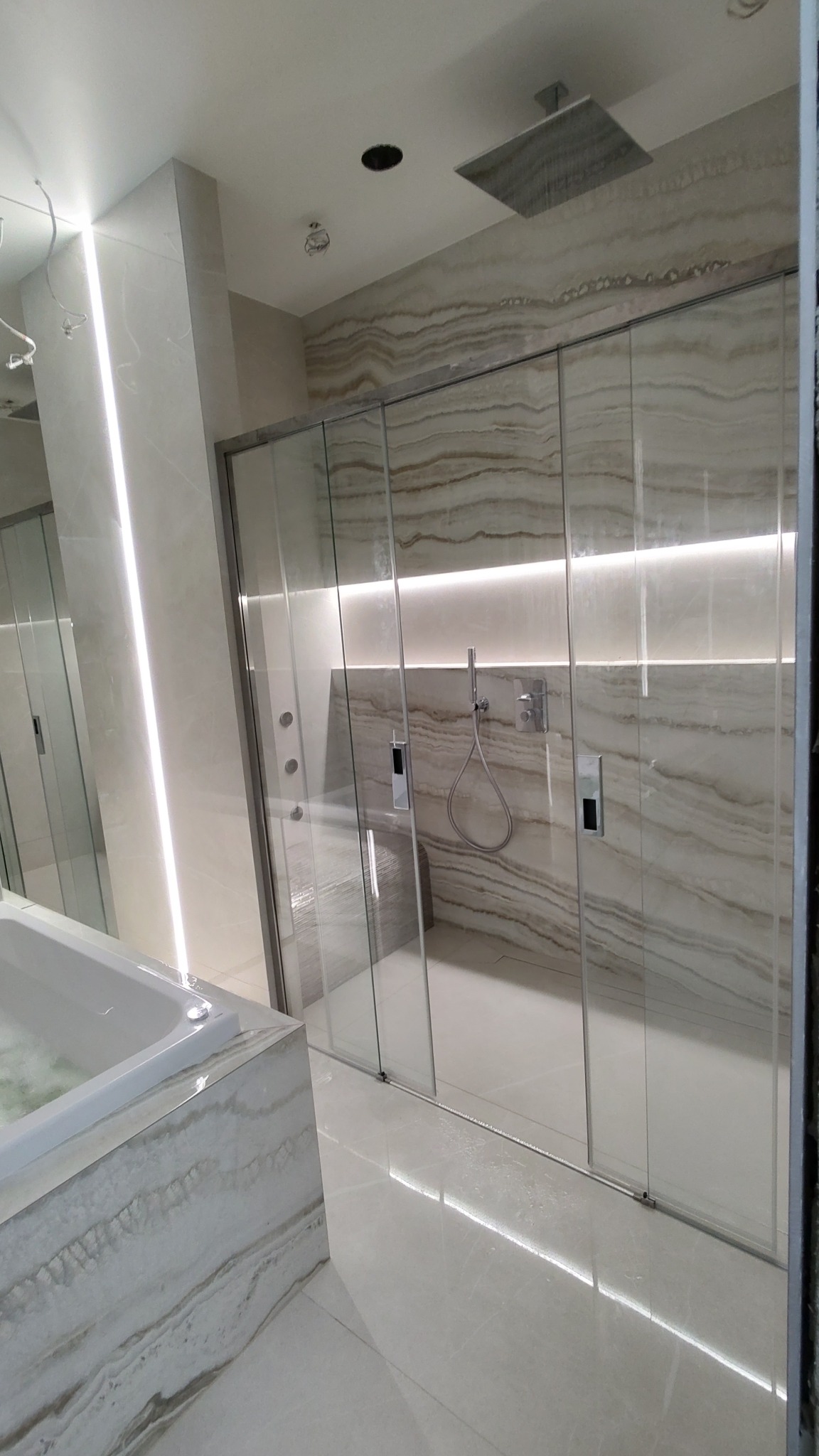 Nowoczesna łazienka z kabiną prysznicową typu walk-in, wykończona płytami imitującymi marmur, z oświetleniem LED wzdłuż ściany i pod prysznicem deszczownicą.