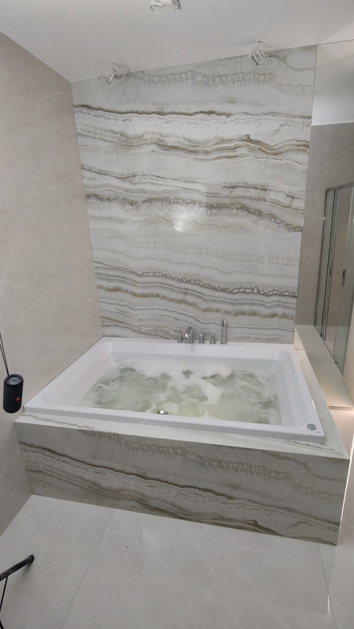 Biała wanna z hydromasażem wypełniona wodą, obłożona płytkami imitującymi marmur, dopasowanymi do płytek ściennych o podobnym wzorze w nowoczesnej łazience.