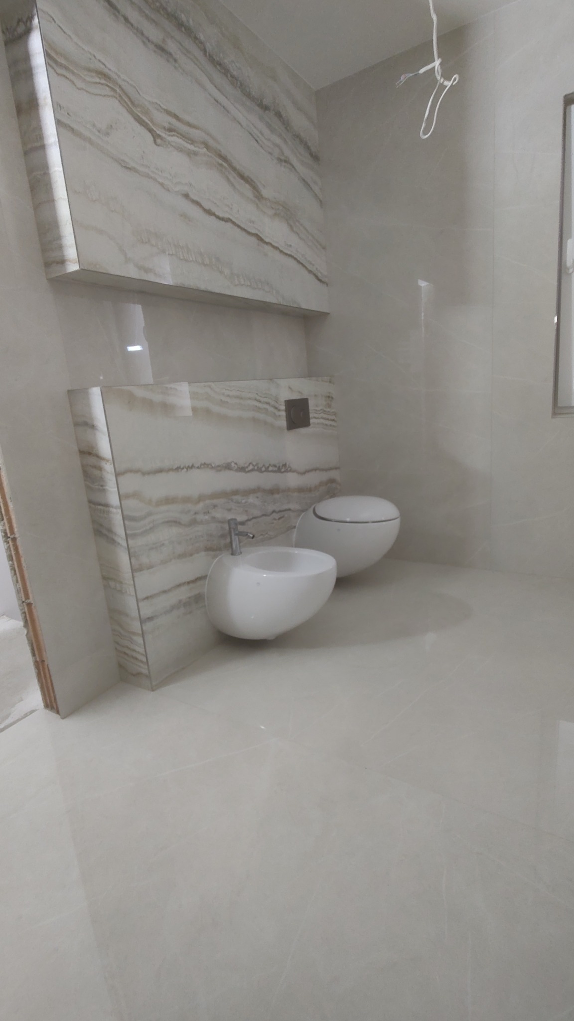 Wykończona łazienka z podwieszaną toaletą i bidetem o zaokrąglonym kształcie, obudowane płytami imitującymi marmur, na jasnych płytkach podłogowych.