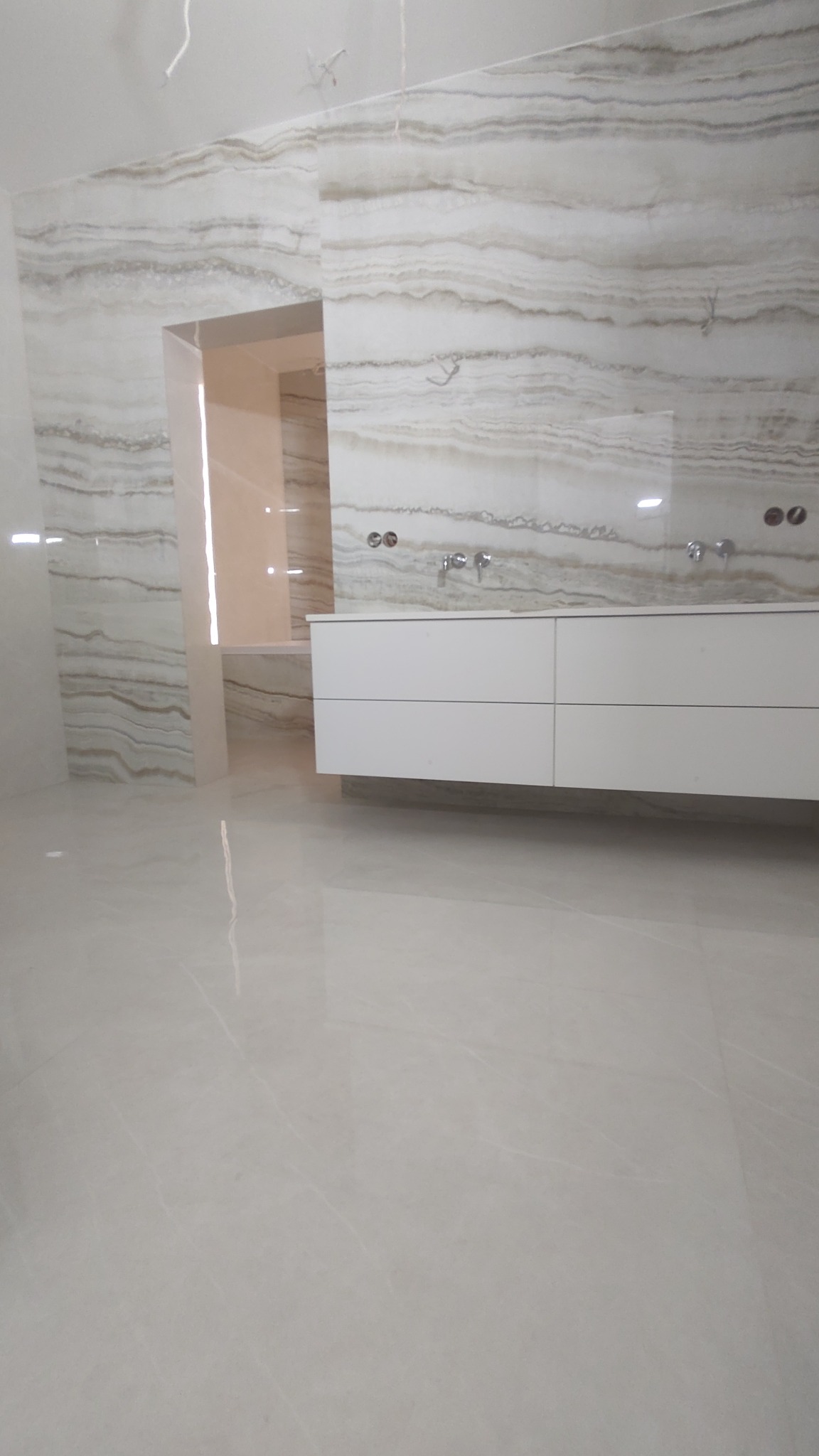 Wykończona łazienka z beżowymi płytkami imitującymi marmur na ścianach, biała, podwieszana szafka pod umywalką z dwoma bateriami, oraz podłoga wyłożona jasnymi płytkami z połyskiem.