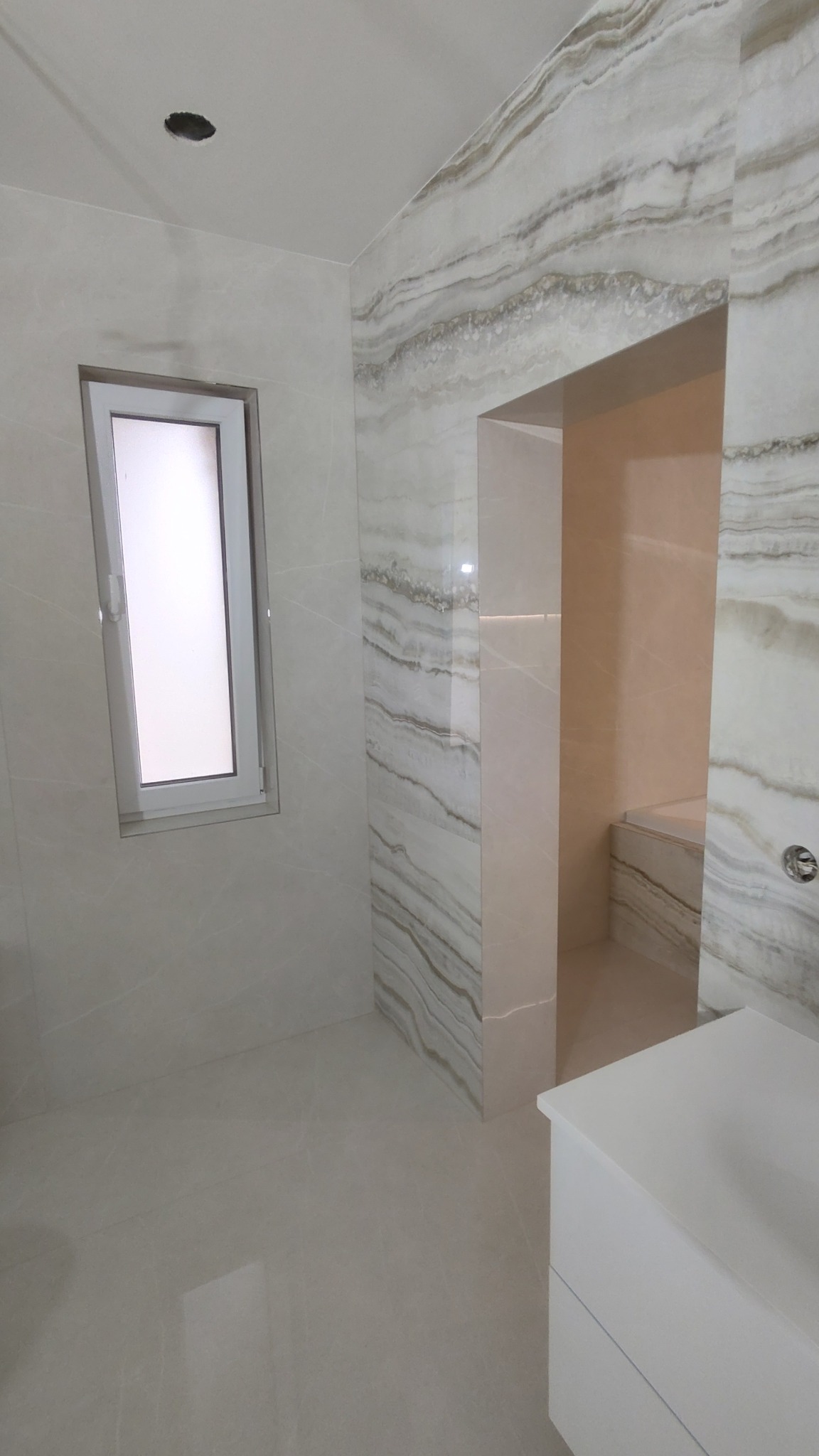 Jasna łazienka z płytkami imitującymi marmur na ścianach, widoczne okno z białymi ramami, fragment szafki umywalkowej i wejście do kabiny prysznicowej.