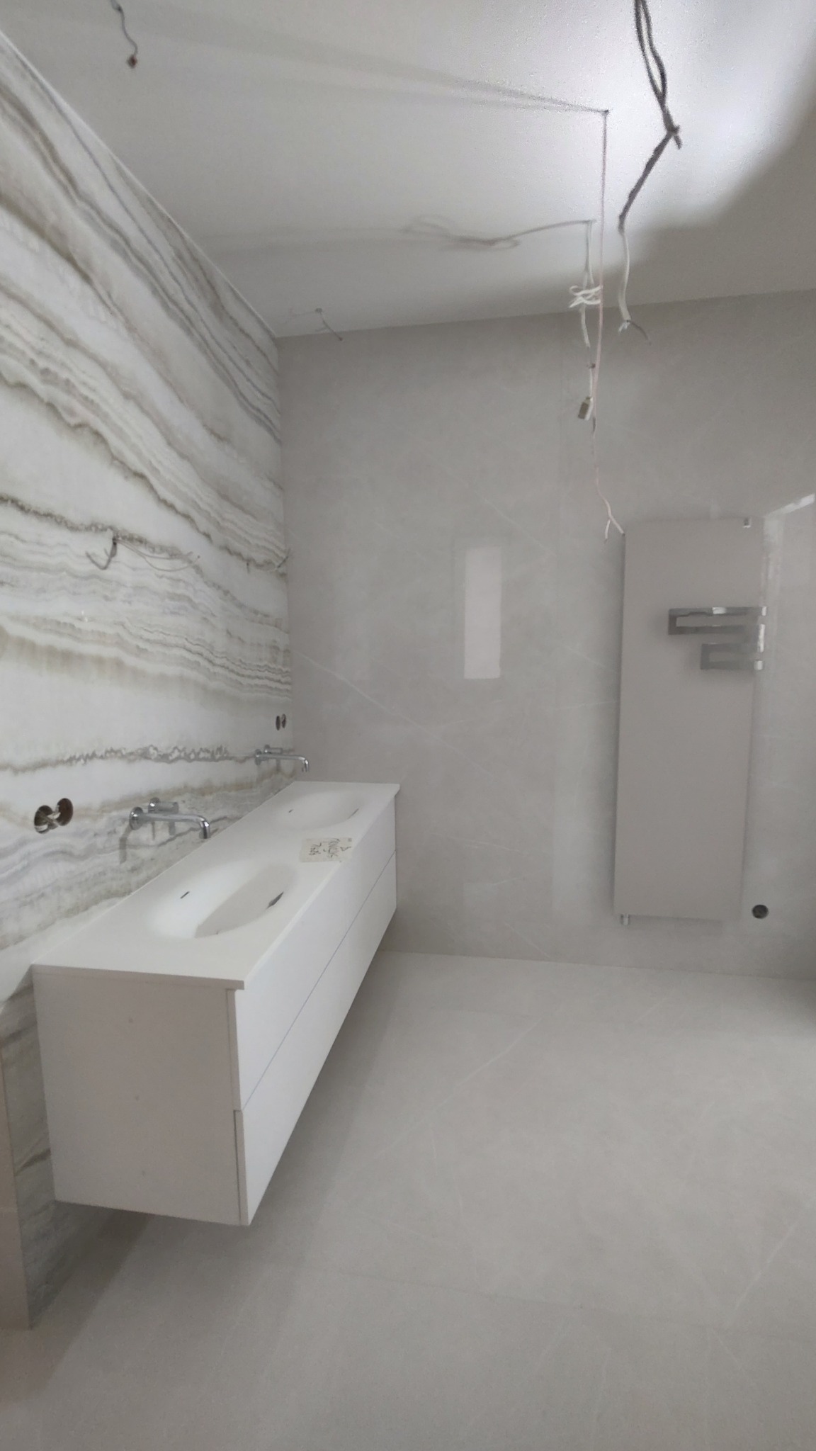 Łazienka w trakcie wykończenia: biała, podwójna umywalka z chromowanymi bateriami na tle ściany z płytek imitujących marmur, obok grzejnik drabinkowy i widoczne przewody elektryczne.
