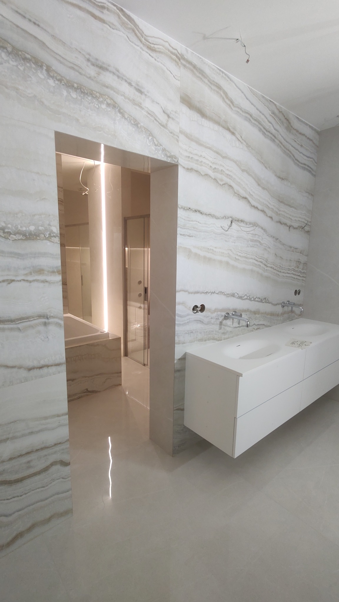 Nowoczesna łazienka z marmurowymi płytami na ścianach i podłodze, biała szafka z umywalkami, widok na kabinę prysznicową i wannę z podświetleniem LED.