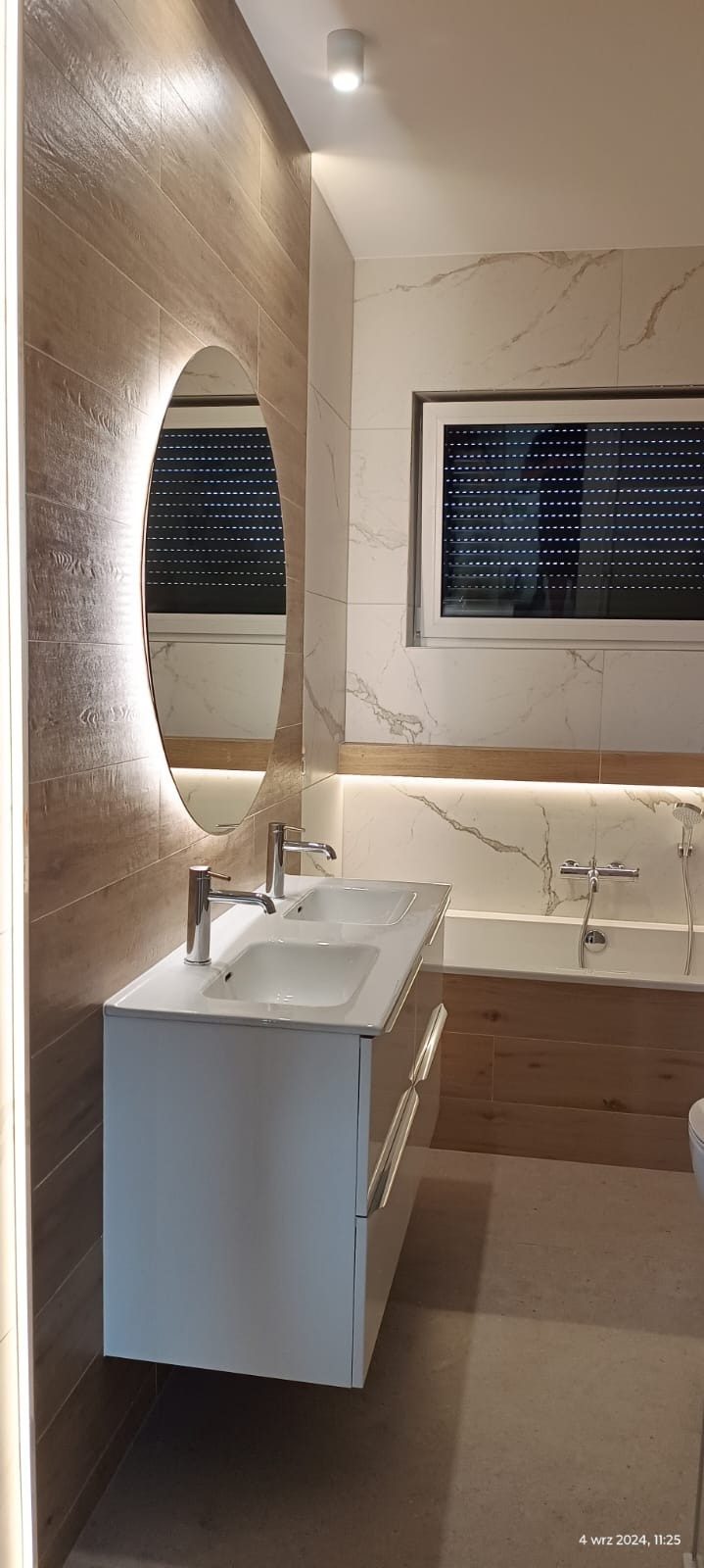 Nowoczesna łazienka z podwójną umywalką na białej szafce, owalnym lustrem z podświetleniem LED, drewnopodobnymi płytkami na ścianie i marmurowymi płytkami nad wanną.