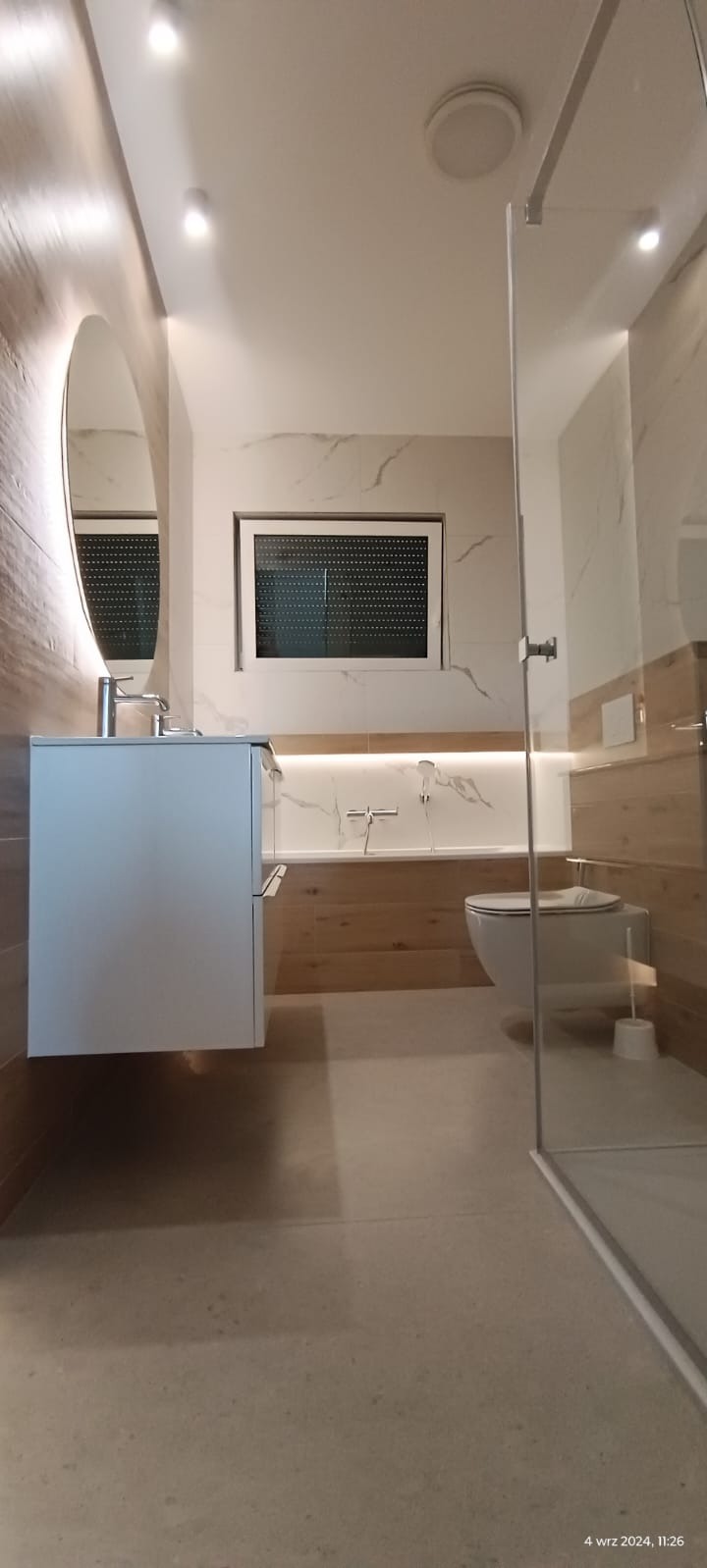Nowoczesna łazienka po remoncie z kabiną prysznicową typu walk-in, umywalką na szafce wiszącej z okrągłym lustrem, oraz oświetleniem LED nad wanną obudowaną płytkami imitującymi drewno.