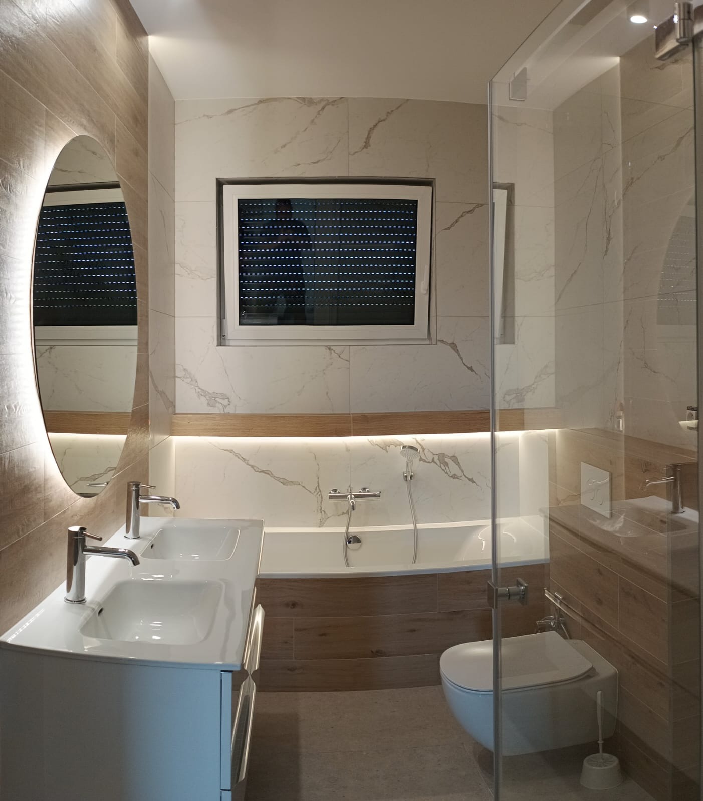Nowoczesna łazienka z podwójną umywalką, owalnym lustrem z podświetleniem LED, wanną, toaletą podwieszaną i oknem z żaluzjami zewnętrznymi.
