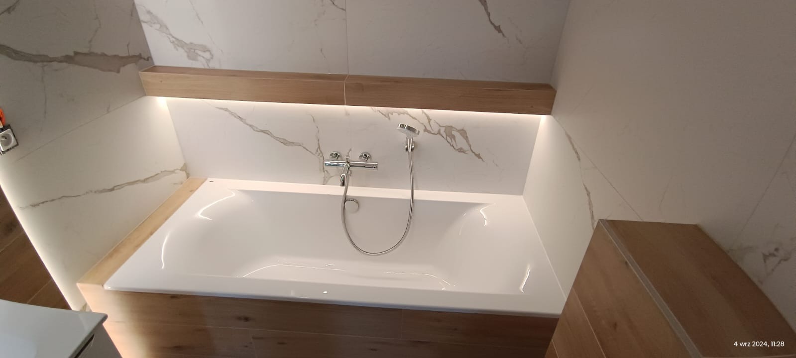 Biała wanna obudowana panelami imitującymi drewno, z chromowaną baterią i słuchawką prysznicową na tle marmurowej ściany z podświetleniem LED.