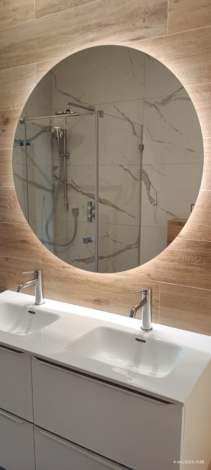 Nowoczesna łazienka z podwójną białą umywalką na szafce, okrągłym lustrem z podświetleniem LED i kabiną prysznicową ze szklanymi drzwiami, wykończona płytkami imitującymi drewno i marmur.
