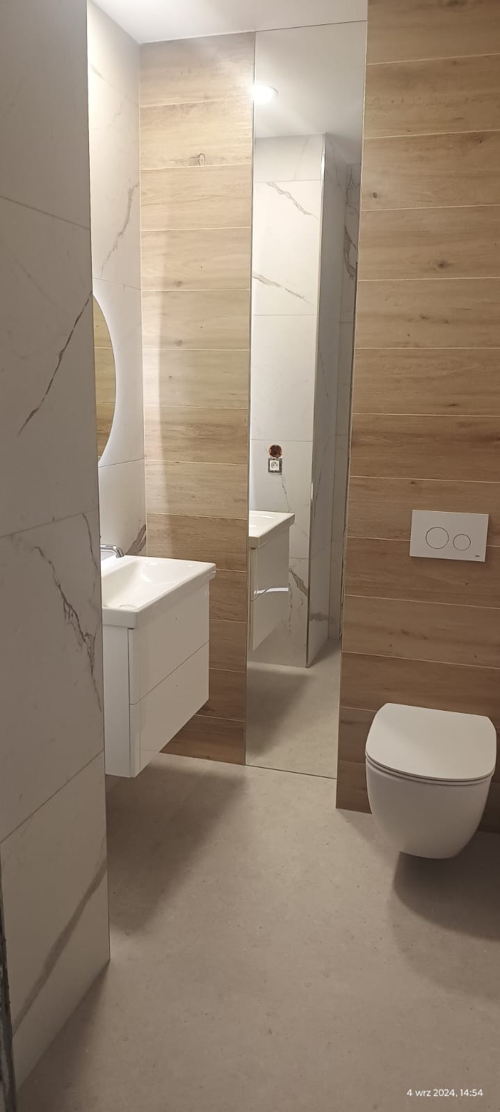 Nowoczesna łazienka z drewnopodobnymi i marmurowymi płytkami, białą umywalką z szafką, okrągłym lustrem i podwieszaną toaletą z przyciskiem dwudzielnym, odbicie częściowo widoczne w lustrzanej płycie.