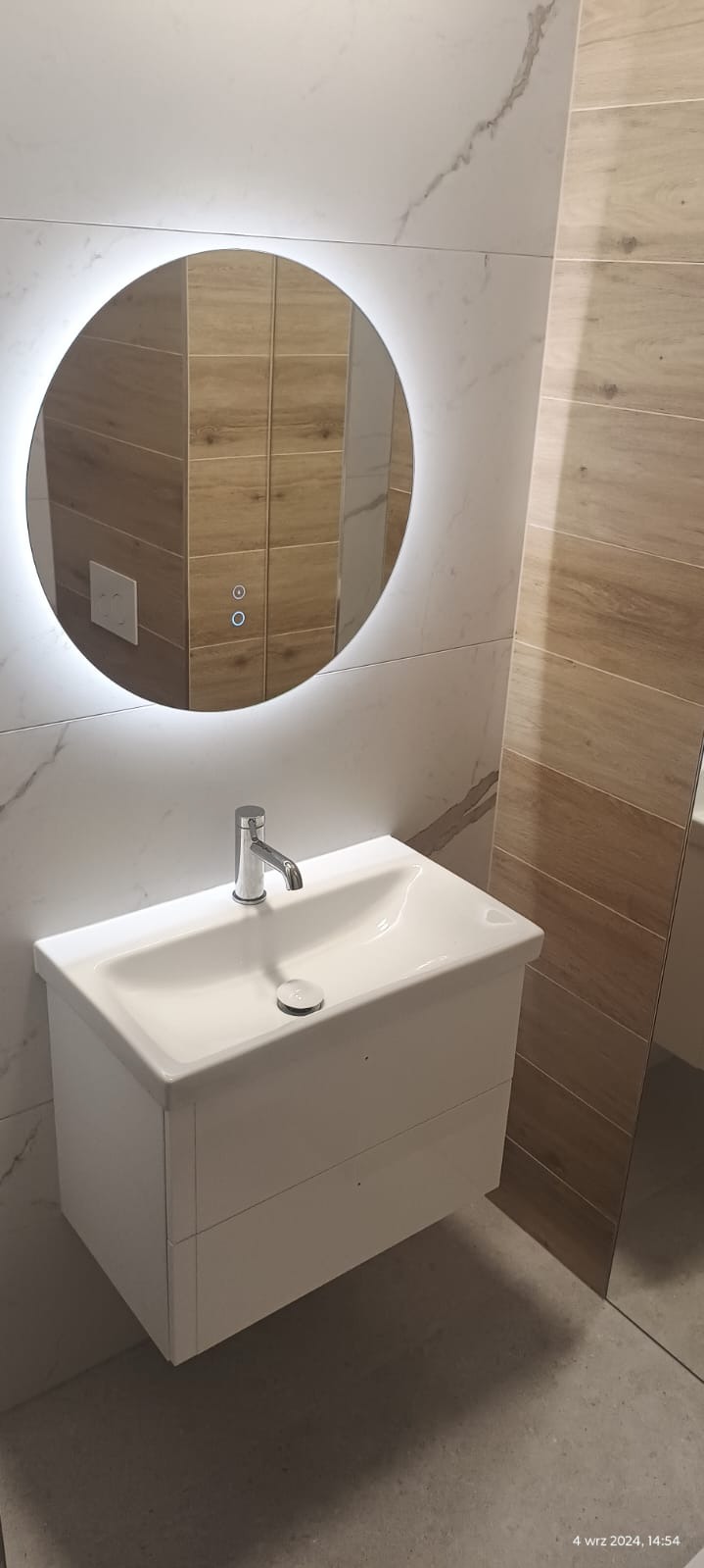 Nowoczesna łazienka z okrągłym lustrem podświetlanym LED, białą umywalką z szafką i armaturą chromowaną, wykończona płytkami imitującymi marmur i drewno.