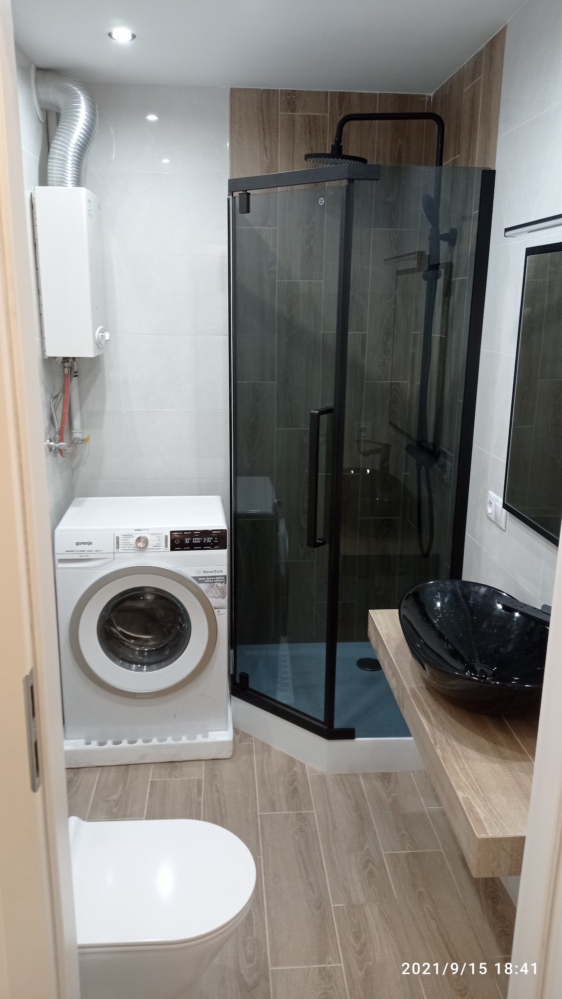 Nowoczesna łazienka po remoncie z kabiną prysznicową z czarnymi ramami, pralką Gorenje, podwieszaną umywalką w kolorze czarnym i grzejnikiem wody, wykończona płytkami imitującymi drewno.