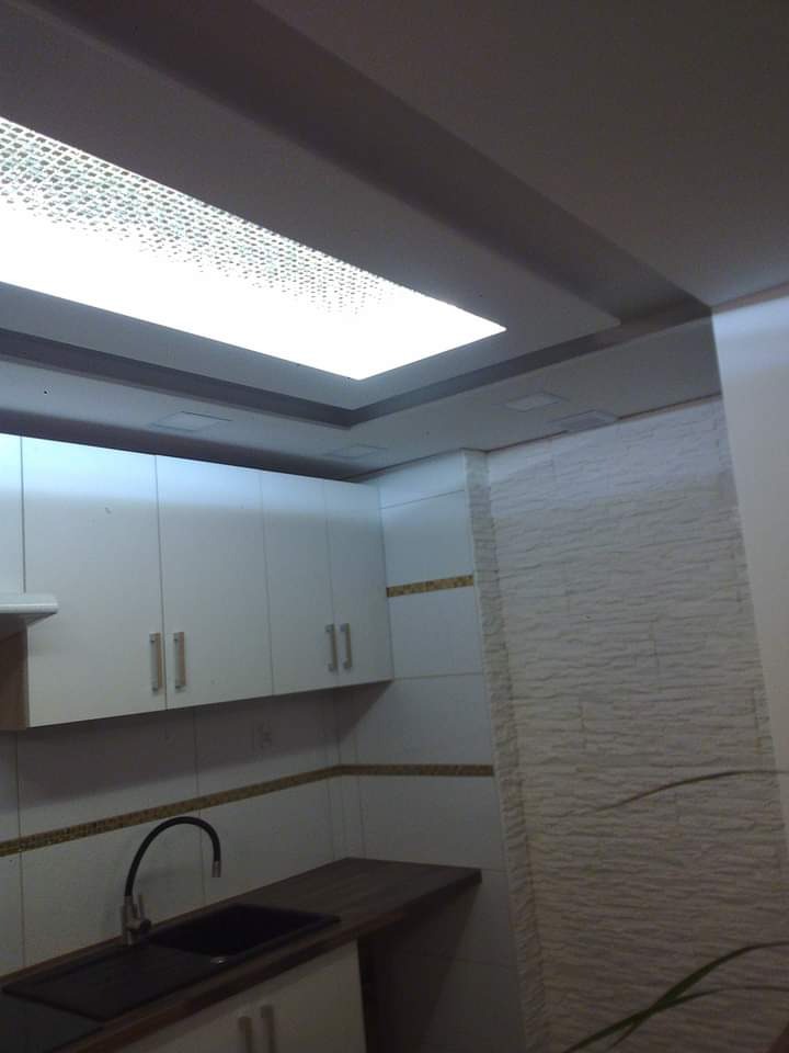 Wykończona kuchnia z białymi szafkami, ciemnym blatem, czarnym zlewozmywakiem i dekoracyjną ścianą z białej cegły, oświetlona nowoczesnym, kasetonowym sufitem.