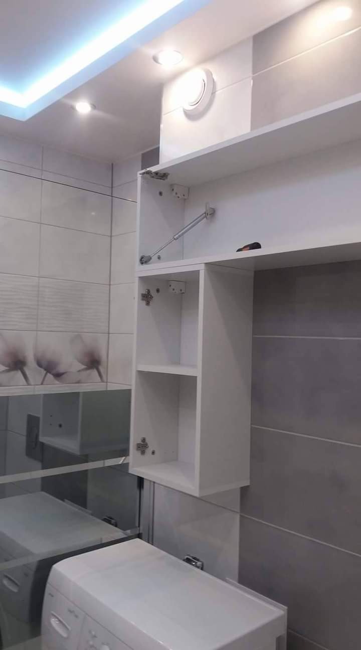 Biała szafka łazienkowa z otwartymi drzwiczkami, widoczny mechanizm podnoszenia, na tle płytek w odcieniach szarości i bieli, obok pralka.
