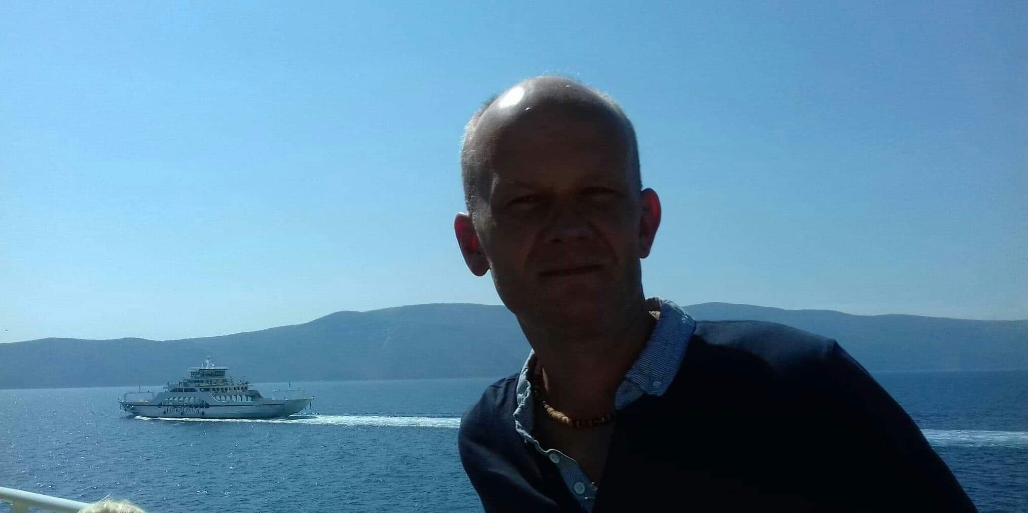 Mężczyzna robiący sobie selfie na tle morza i przepływającego promu w słoneczny dzień.