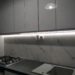 Biała kuchnia z połyskiem, marmurowe płytki na ścianie, czarny blat, oświetlenie LED pod szafkami, widoczna płyta gazowa i zlew.