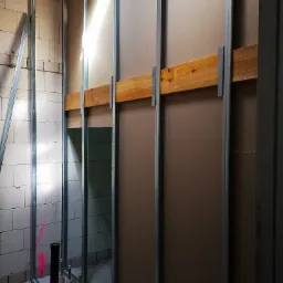 Wnętrze w trakcie budowy: metalowy szkielet ścian działowych z zamontowaną częściowo płytą kartonowo-gipsową, widoczne drewniane wzmocnienia i ślady różowej farby na ścianie z bloczków.