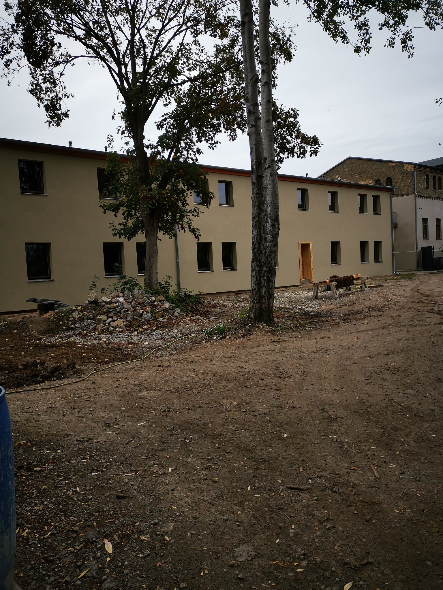 Dwukondygnacyjny budynek z nową elewacją w kolorze beżowym, widoczne nowe okna i surowe drzwi, teren wokół budynku w trakcie prac ziemnych, drzewa na pierwszym planie.